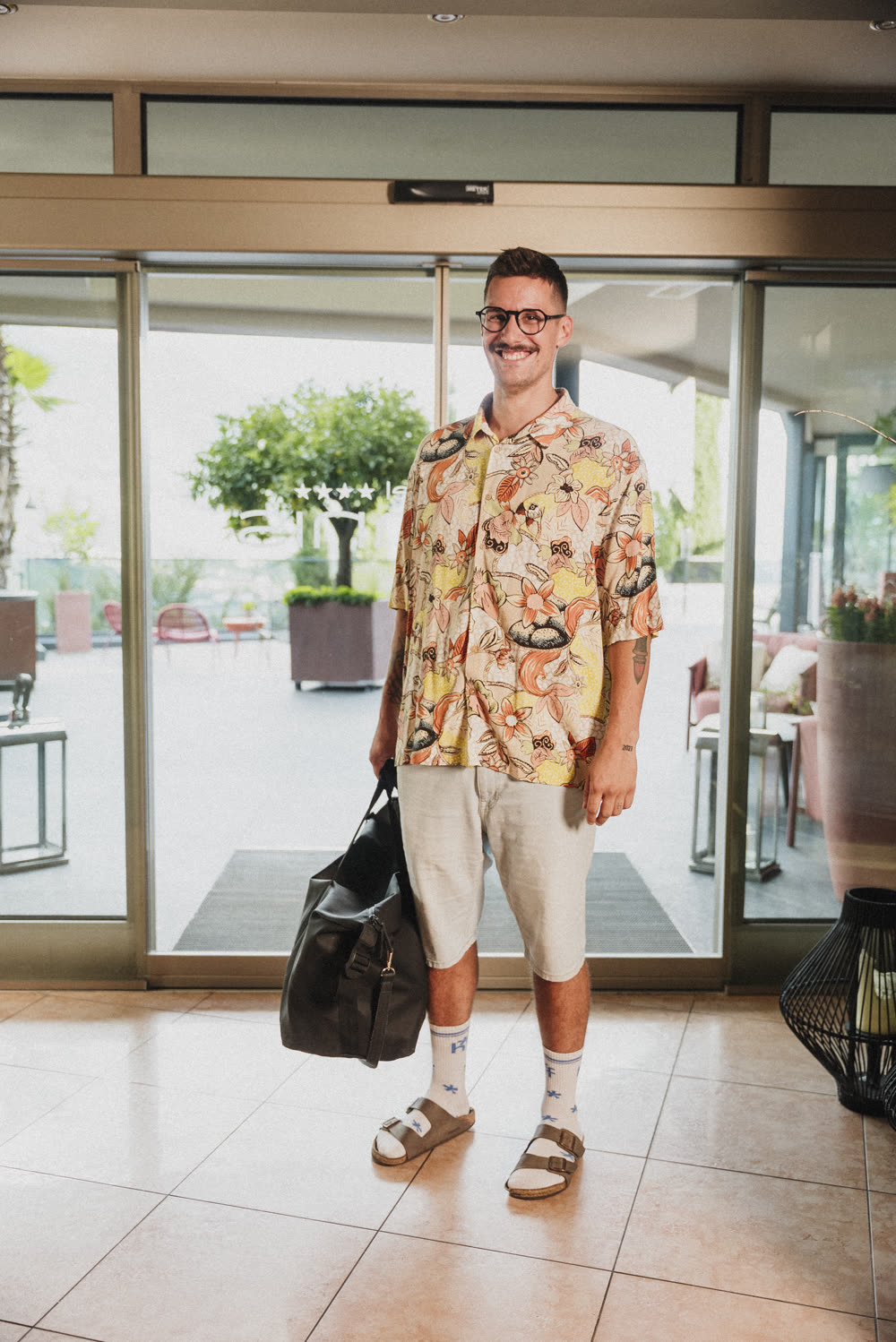 A guest in a Hawaiian shirt walks through the entrance door with a travel bag